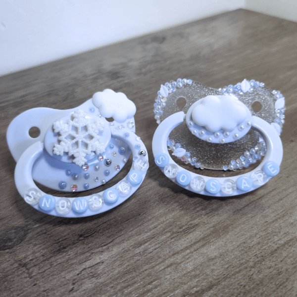 a silver and white snow themed paci and white paci with cloud and snowflake decorations